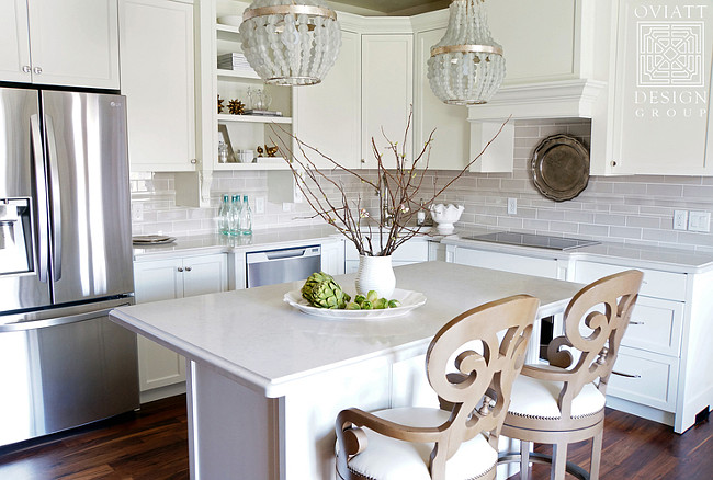 Small Kitchen Reno. The cabinet color is Restoration Hardware Cloud White and the wall color is 1/2 strength BM Edgecomb Gray. A pair of gray beaded chandeliers hangs over a small kitchen island lined with Noir Saragossa Swivel Stools. Creamy white shaker cabinets are paired with ivory quartz countertops and a gray stacked tile backsplash. A corner kitchen sink is flanked by a dishwasher to the left and an induction cooktop to the right. #Kitchen #KItchenREno #Reno #KitchenProject Oviatt Design Group.