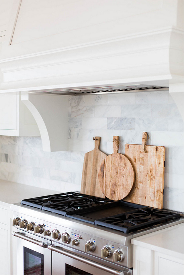 White Marble Brick Backsplash. A white kitchen hood with corbels stands over a white and grey marble brick tiled backsplash and a stainless steel stove lined with cutting boards. #MarbleBacksplash #KitchenBacksplash #Backsplash #KitchenMarbleBacksplash Ashley Winn Design. 