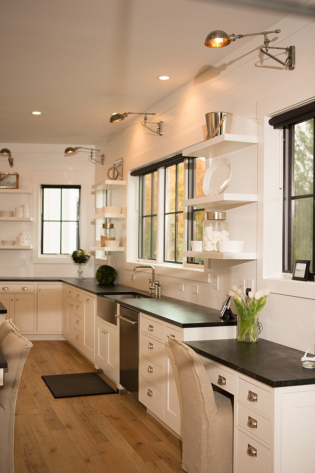 Kitchen Desk. Soapstone kitchen desk. This kitchen features shiplap walls lined with white cabinets paired with soapstone countertops fitted with a stainless steel apron sink under a window flanked by stacked floating shelves. Kitchen with a drop down soapstone top desk placed under a window illuminated by a swing arm sconce. #Kitchen #Desk #KitchenDesk Hahn Builders.