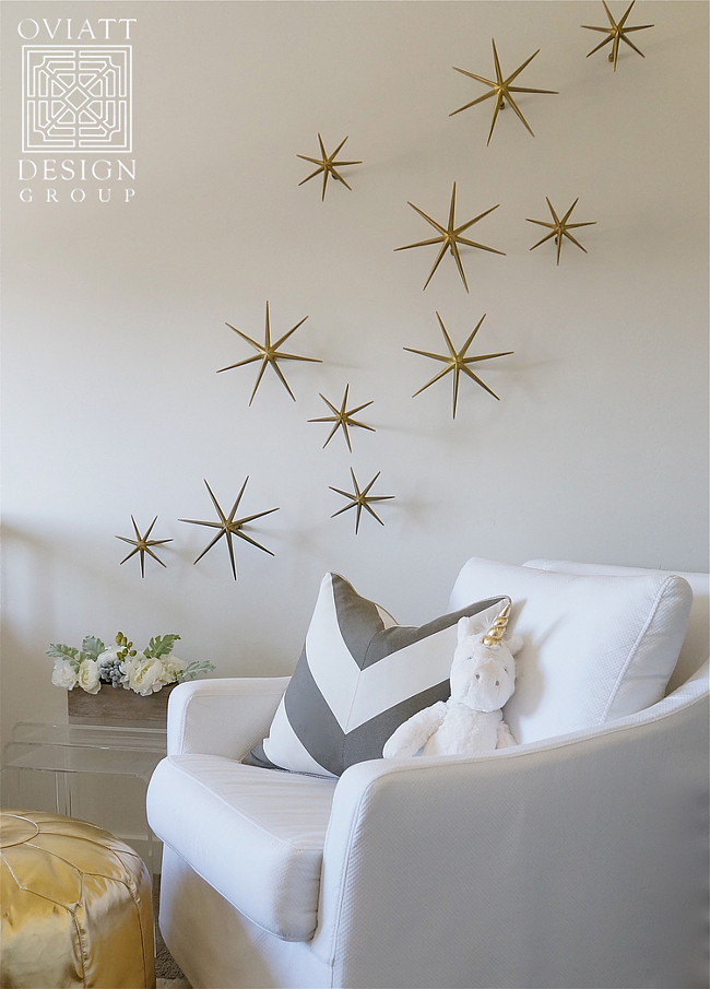 Neutral Gender Nursery. White and gold nursery boasts a white slipcovered glider lined with a gray chevron pillow next to a gold Moroccan pouf placed under gold wall stars, Global Views Etoile Brass Wall Décor. #Neutral #Genger #Nursery Oviatt Design Group.