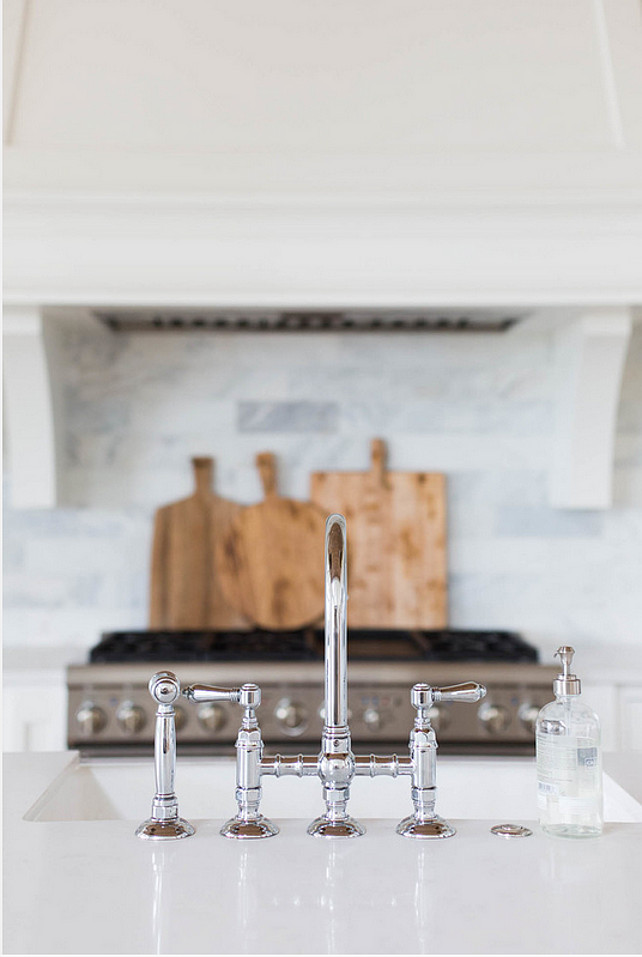 Kitchen Faucet. Kitchen island topped with white quartz fitted with a farmhouse sink and deck mount faucet. A white kitchen hood with corbels stands over a marble brick tiled backsplash and a stainless steel stove. The kitchen faucet is the Rohl Polished Nickel Country Kitchen Three Leg Bridge Faucet. #KitchenIsland #KitchenFaucet #Faucet #Kitchen Ashley Winn Design