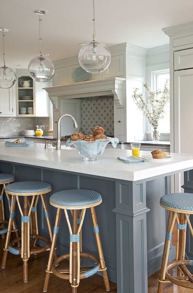 Two toned kitchen with blue island. kitchen with clear glass globe pendants over blue kitchen island with white marble countertops and sink lined with blue French cafe bar stools. Two-tone kitchen features light gray cabinets paired with black countertops and linear light gray tile backsplash. Light gray kitchen hood features pull-out spice racks flanking cooktop as well as corner open display cabinet. #Kitchen #TwotonedKitchen Kerry Hanson Design.