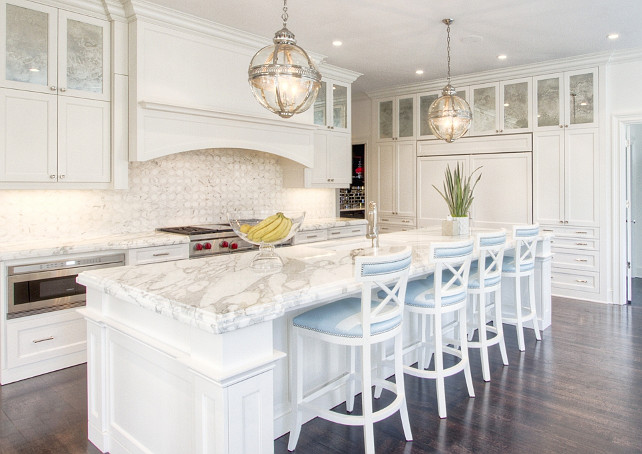 Classic White Kitchen. Classic white kitchen with antique mirror cabinets. Exquisite kitchen features a pair of Restoration Hardware Victoria Hotel Pendants illuminating an super long center island topped with white marble framing square sink and polished nickel faucet lined with white and blue counter stools. A curved kitchen hood stands over a mosaic marble backsplash and a Wolf Range with an under the counter microwave to the left. The back wall of the kitchen is fitted with antiqued mirrored cabinets stacked over pantry cabinets flanking a paneled refrigerator and freezer. Anne Hepfer Designs.