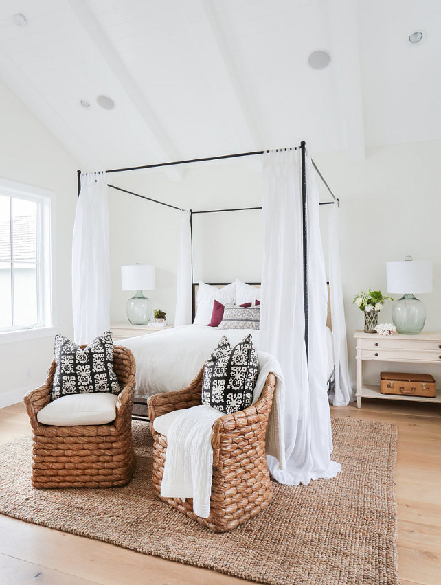 This beach-y bedroom features a vaulted ceiling with white wood beams over an iron canopy bed. The whitewashed wood farmhouse nightstands are decorated with recycled glass lamps and fresh flowers. Notice the wide plank floors, the seagrass chairs with black and white pillows and the jute rug. 