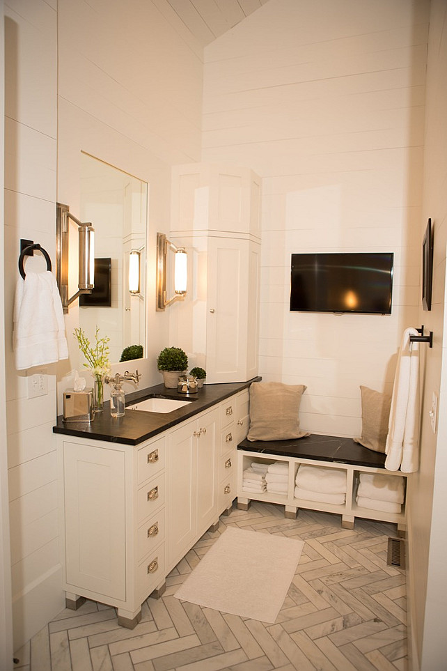 Master Bathroom Ideas. Master bathroom features a white washstand topped with soapstone under an inset mirror illuminated by Eclipse Wall Lights next to a corner cabinet and a built-in soapstone bench with open cubbies filled with fluffy white towels situated under a flatscreen TV alongside a marble herringbone floor. #MasterBathroom #Bathroom Hahn Builders.