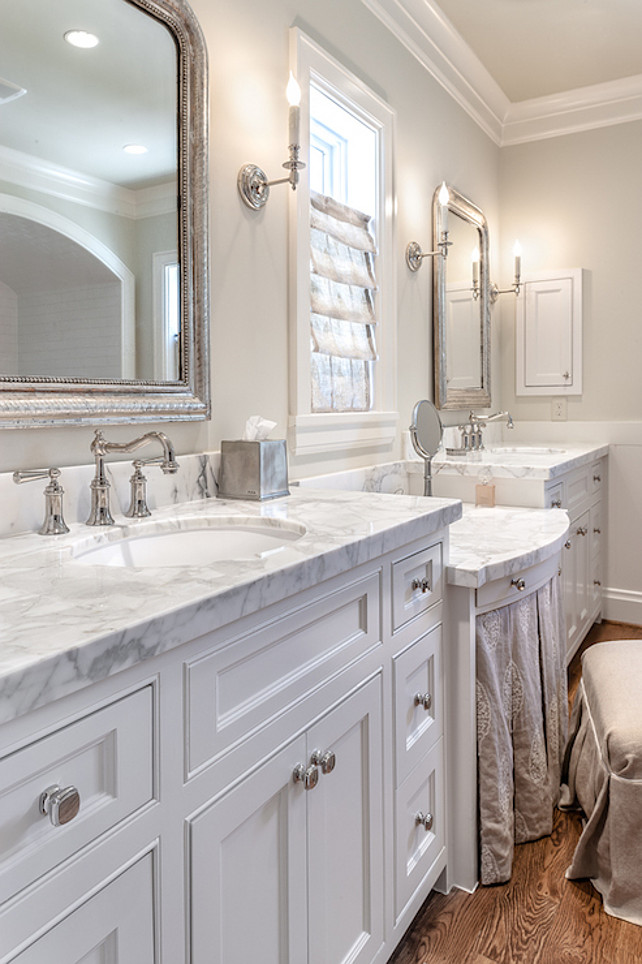 Bathroom Cabinet Ideas. Bathroom features light gray walls adorned with crisp white crown molding and base boards framing a drop-down skirted make up vanity topped with white and gray marble paired with gray linen vanity bench situated under window dressed in gray roman shade illuminated by candlestick sconces flanked by his and her sink cabinets paired with marble countertops under Eloquence Louis Philippe Mirrors. #bathroom #Bathroom Cabinet Frasier Homes