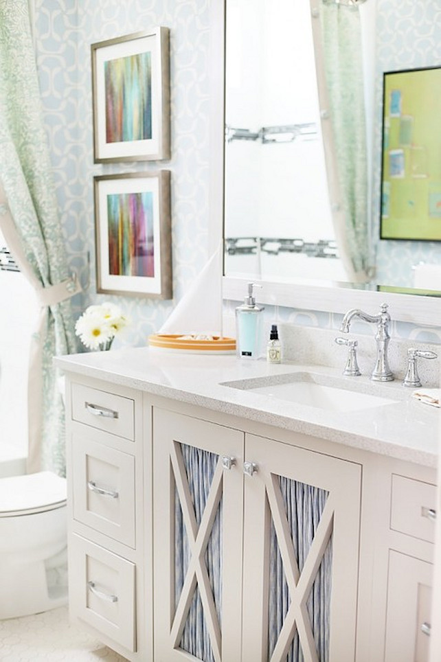 Bathroom. Pretty bathroom features white and blue geometric wallpaper on walls which highlight a pair of framed abstracts hung over the toilet to the left of an ivory sink vanity with x-front cut out cabinet doors dressed in blue fabric below an ivory Corian countertop which frames an undermount porcelain sink and hook spout faucet below a matching ivory mirror. #bathroom UV Parade of Homes 