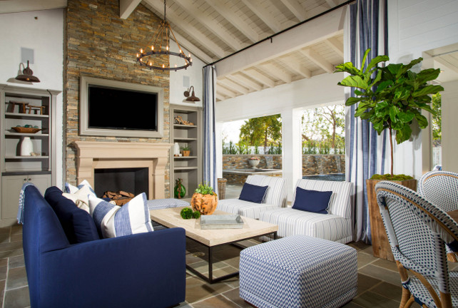 Family room. Coastal blue and white family room. The living room opens to an outdoor living area that features a TV on stone fireplace flanked by gray built-in bookcases illuminated by oil-rubbed bronze vintage lanterns across from blue sofa facing white and gray striped slipper chairs accented with navy blue pillows across from industrial coffee table atop slate tiled floor. Interiors Legacy Custom Homes, Inc.