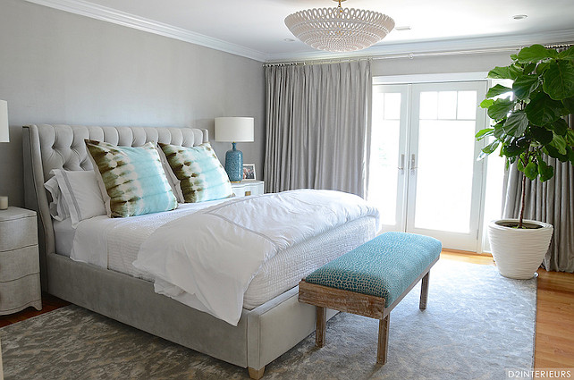 Gray Bedroom. Gray Bedroom Design. Stunning gray bedroom accented with pops of turquoise blue features gray walls alongside French doors dressed in gray silk drapes illuminated by an Oly Studio Pipa Bowl Chandelier. A weathered bench with turquoise cushion stands at the foot of the gray velvet bed with tufted headboard which is dressed in white and gray hotel bedding and aqua blue tie dye pillows flanked by Studio A Toile Linen Chests topped with AERIN Culloden Table Lamps atop a gray ikat rug.