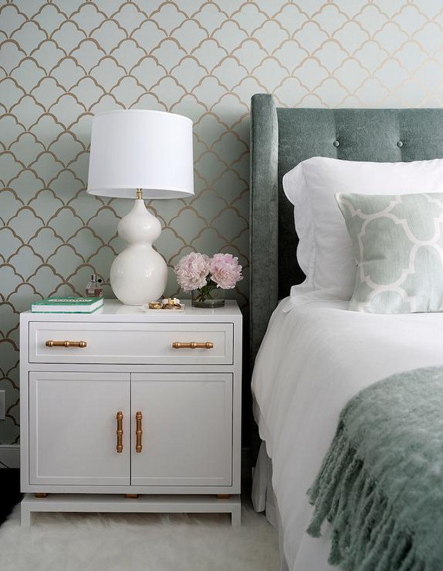 Master Bedroom Bedding Ideas. Grey bedroom features an accent wall clad in green and gold scallops wallpaper lined with a gray velvet wingback headboard on bed dressed in white scalloped bedding, green Moroccan tiles pillows in Windsor Smith Riad Fabric and a gray fringed throw blanket. A gray tufted headboard stands next to a white nightstand with gold bamboo pulls, Worlds Away Marcus White & Gold Leaf Cabinet, topped with a white glass lamp atop a white sheepskin rug.