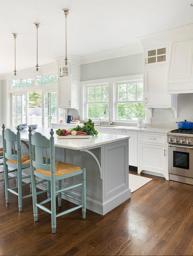 White and Gray Kitchen Painted in Benjamin Moore OC-52 Gray Owl. In this kitchen, a white paneled kitchen hood stands over the backsplash and the stainless steel stove, right next to a sink with gooseneck faucet. Notice the corbels on the island and the upper cabinets. Floors are oak. Digs Design Company.