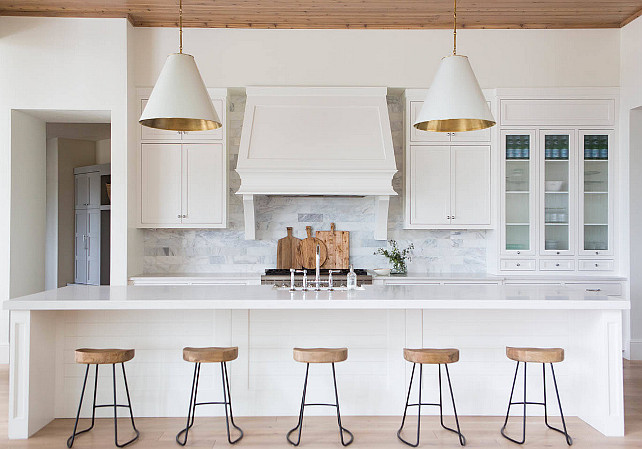 Transitional Kitchen. Transitional White Kitchen. A pair of white and gold cone pendants, Goodman Hanging Lamps, illuminates an extra-long center island topped with white quartz fitted with a farmhouse sink and deck mount faucet lined with wood and iron counter stools, Wisteria Smart & Sleek Stools. A white kitchen hood with corbels stands over a marble brick tiled backsplash and a stainless steel stove. #Kitchen Ashley Winn Design.