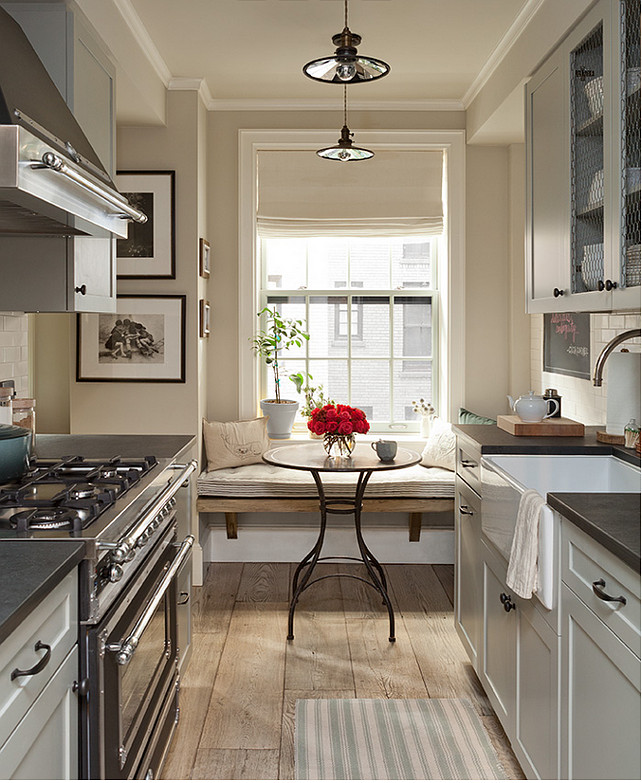 Galley Kitchen. Small Galley Kitchen. Grey Galley Kitchen. Small galley kitchen features upper cabinets with chicken wire doors over light gray shaker cabinets paired with honed black countertops framing a farmhouse sink accented with a gooseneck faucet and a mini subway tiled backsplash situated across from a stainless steel range hood over a black stove atop rustic wide plank floors adorned with a white and green striped rug. The back of the galley kitchen boasts a breakfast nook filled with a rustic bench doubling as window seat facing a round wood and iron bistro table illuminated by vintage barn pendants. Jenny Wolf Interiors.