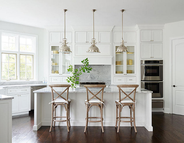 White Kitchen. Classic White Kitchen. White Kitchen kitchen features three Restoration Hardware Harmon Pendants illuminating a white center island topped with gray quartzite lined with three French x back counter stools. A kitchen hood, flanked by glass front cabinets filled with yellow dinnerware, stands over a gray quartzite backsplash and a stainless steel stove. Kitchen with double wall ovens stacked above a stainless steel warming drawer next to a corner walk in pantry. #Kitchen #WhiteKitchen Shophouse Design.