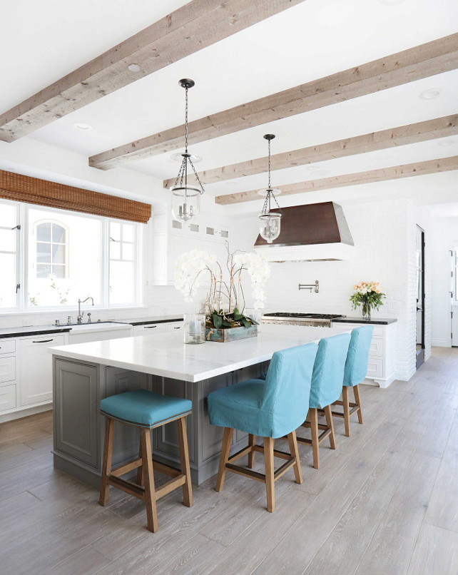 Kitchen Grey Wood Floors. Kitchen with White Cabinets & Gray Island. White kitchen boasts a ceiling clad in wood beams and glass lanterns illuminating a gray center island topped with white marble lined with turquoise blue slipcovered counter stools atop a gray wood floor. A black and white kitchen hood stands over a swing arm pot filler and a high end stainless steel stove. Kitchen with white cabinets paired with black marble countertops and a white brick wall backsplash. A farmhouse sink and a vintage style faucet is placed below windows dressed in one bamboo roman shade. #Kitchen #KitchenFloors #GreyFloors Blackband Design.