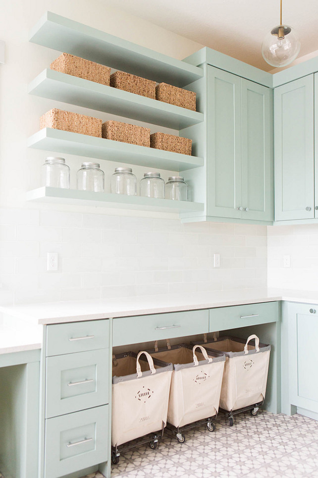 Laundry Room Design. Laundry room features gray green cabinets paired with white quartz countertops and a white glass subway tiled backsplash. Laundry room with three Steele Canvas laundry carts tucked below a countertop placed under stacked gray green floating shelves filled with woven baskets and vintage jars illuminated by a brass and glass globe pendant. #LaundryRoom Ashley Winn Design. 