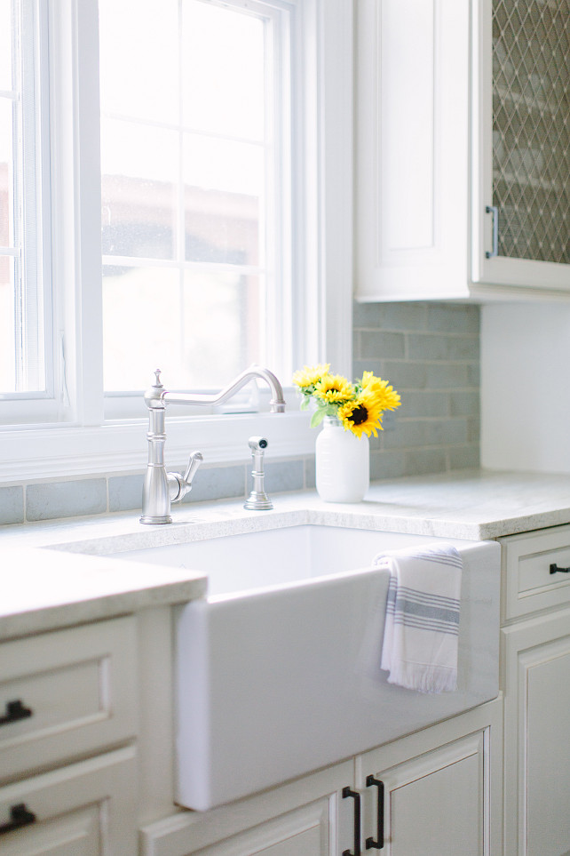 Farmhouse Sink. Kitchen with white farmhouse sink. Kitchen features white raised panel cabinets adorned with oil rubbed bronze pulls paired with white marble countertops and a blue beveled subway tiled backsplash. A farmhouse sink stands under a hook and spout faucet placed under windows flanked by cabinets fitted with metal lattice doors. #Kitchen #FarmhouseSink #Sink #KitchenSink #ApronSink Kate Marker Interiors.