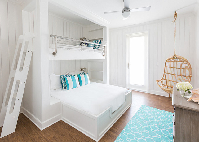 Bunk room with a built-in bed situated under a loft bed with safety rails, both dressed in soft white bedding and turquoise pillows, and fitted with modern ladder situated across from a gray wash dresser alongside a turquoise hex rug next to a Two's Company Hanging Rattan Chair. Laura U, Inc. 