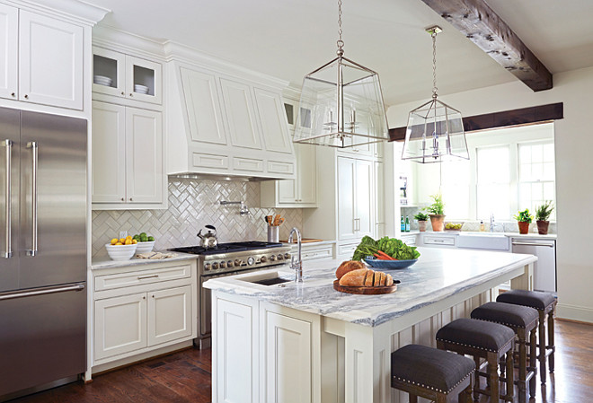 Kitchen kitchen features white shaker cabinets paired with marble countertops and a white herringbone pattern backsplash. A white paneled hood over a swing-arm pot filler and high-end stainless steel stove flanked by an under cabinet stainless steel refrigerator to the left and full height pantry cabinets to the right. A pair of glass and nickel lanterns illuminate a paneled kitchen island topped with white and gray marble framing a corner prep sink paired with gooseneck faucet lined with backless brown nailhead counter stools with silver nailhead trim. The back of the kitchen boasts a nook filled with a farmhouse sink next to stainless steel dishwasher under three windows flanked by built-in shelving. Birmingham Home and Garden. 