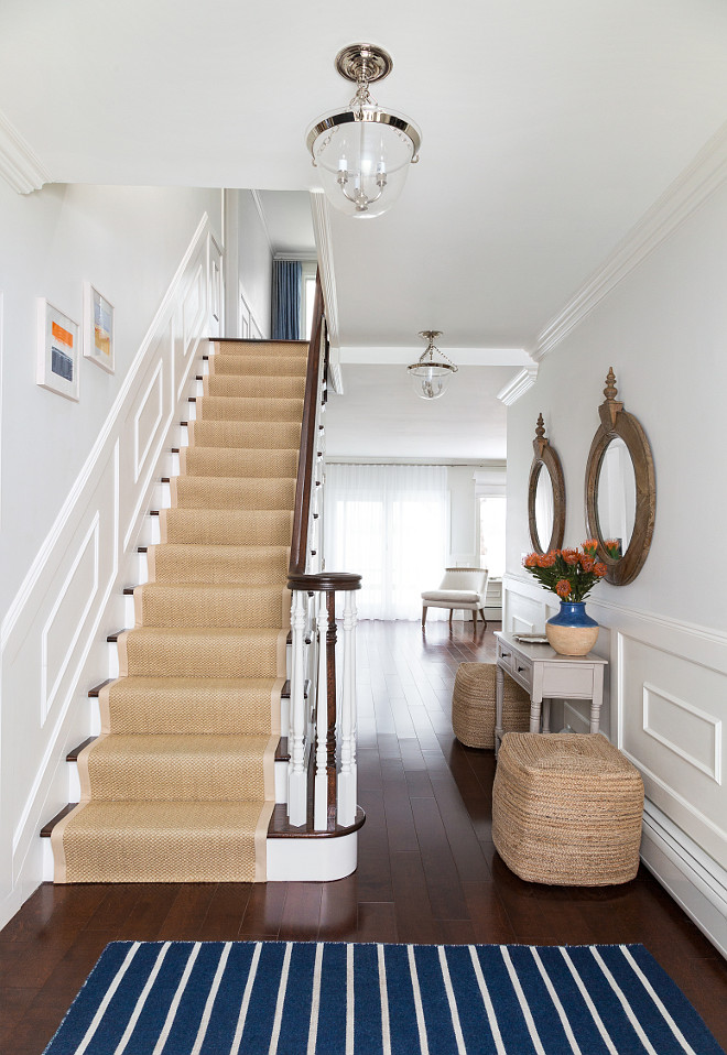 White Foyer with Blue Striped Rug. Paint Color is BM White Dove. Narrow foyer features white paint on upper wall and wainscoting on lower wall lined with side by side wood mirrors over a gray console table and a pair of seagrass cube poufs. Cottage foyer boasts a traditional staircase clad in wainscoting lined with a bound sisal runner illuminated by a glass bell jar lantern. #Foyer #WhiteFoyer #BlueStripedRug Chango & Co.