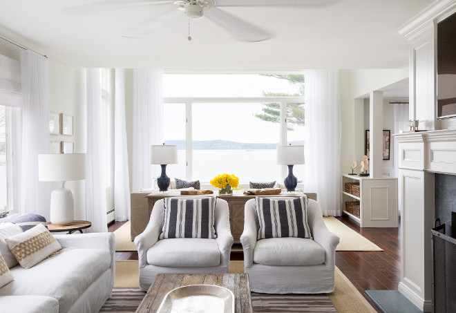 Living Room Decor Ideas. The living space boasts a white sofa flanked by round industrial end tables next to a pair of white slipcovered chairs adorned with blue and grey striped pillows facing a wood and chrome bench coffee table atop a striped rug layered over a bound sisal rug. #LivingRoom #Decor #Interiors Chango & Co. 