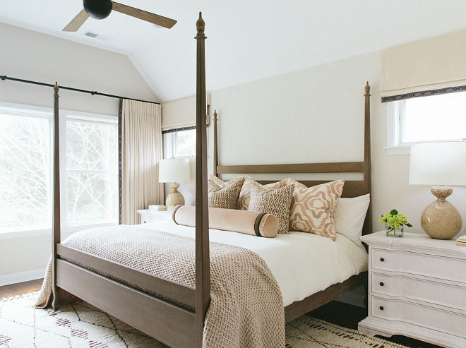 Neutral bedroom features a brown 4 poster bed dressed in pink and brown pillows as well as a pink and brown velvet bolster pillow flanked by whitewashed nightstands and brown lamps, Aerin Gannet Lamps, placed under windows dressed in cream roman shades. Kate Marker Interiors 