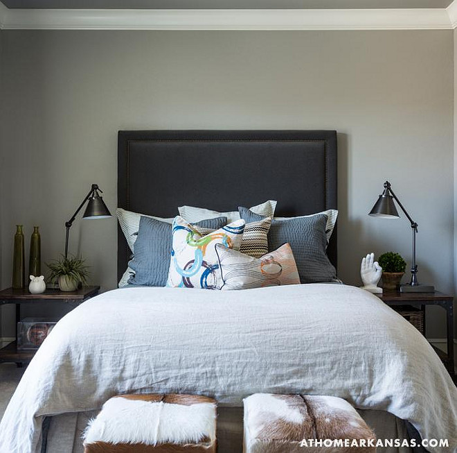 Masculine bedroom with gray walls highlighting a charcoal gray velvet headboard on queen bed layered with linen euro shams and gray matelasse pillows with a cluster of abstract pillows in front flanked by iron and wood nightstands topped with adjustable bronze task lamps with a pair of cowhide cubes at the foot of the bed. At Home in Arkansas.