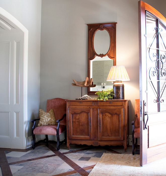 Farmhouse Foyer. Farmhouse foyer with antler arched door filagree footed commode footed console interior light gray walls pink upholstery recessed panel cabinet doors table lamp tile floor wainscoting wood mirror frame wrought iron #Farmhouse #FarmhouseInteriors #Foyer Allard Ward Architects.