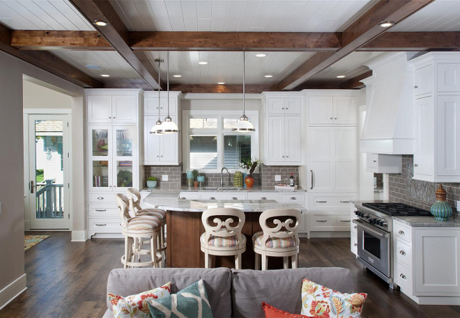 White kitchen with gray backsplash. Gray backsplash tiles. In the kitchen, crisp white cabinetry by Benchmark Wood Studio joins subway tile and granite countertops for a timeless touch. The island, which matches the alder beams on the ceiling, comfortably seats up to five people in the light and airy space. White kicthen gray backsplash ideas. #Whitekicthen #graybacksplash 