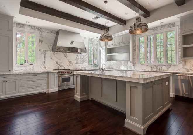 White and gray kitchen features a tray ceiling lined with dark stained wood beams and a pair of Currey and Company Syllabus Pendants illuminating a gray center island fitted gray and white marble countertop framing a corner prep sink situated across from gray open shelving flanking window. Gray kitchen cabinets are paired with gray and white marble countertops and matching backsplash. A stainless steel kitchen hood flanked by windows stands over a Wolf Double Range atop cherry stained floors. Elizabeth Garrett Interiors