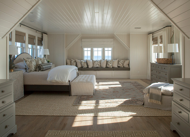 Bedroom window seat. Cream and gray bedroom features a cream beadboard ceiling over a gray bed with a cream arched headboard dressed in cream and gray bedding flanked by gray faux bois skirted bedside tables in Duralee Philip Gorrivan London Plane Fabric and gray lamps, Large Narrow Neck Vase Form, placed in front of windows dressed in bamboo roman shades. A pin stripe tufted bench is placed at the foot of the bed atop a blue and brown Persian rug layered atop a Greek key jute rug facing a gray dresser and Barbara Barry Simple Scallop Table Lamps. Chic master bedroom a window seat alcove fitted with a bench lined with pillows. #Bedroomwindowseat #bedroom #windowseat #bedrooms #Masterbedroom #windowalcove Urban Grace Interiors.