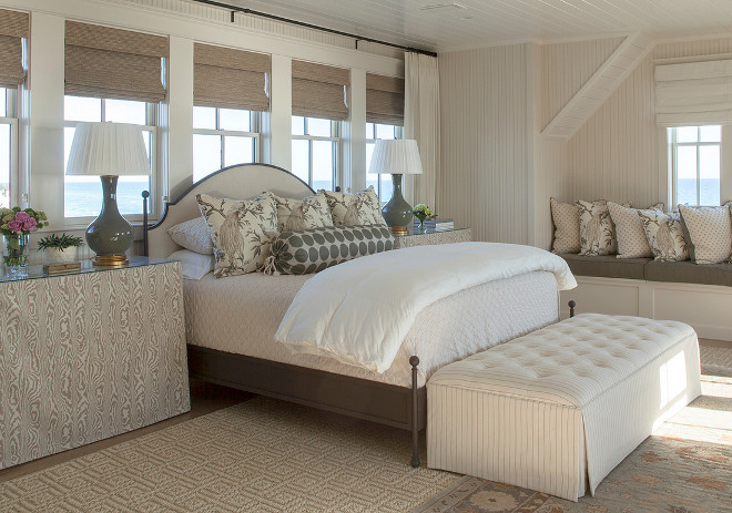 Ivory bedroom features a cream beadboard ceiling over a gray bed with a cream arched headboard dressed in cream and gray bedding flanked by gray faux bois skirted bedside tables in Duralee Philip Gorrivan London Plane Fabric and gray lamps, Large Narrow Neck Vase Form, placed in front of windows dressed in bamboo roman shades. A pin stripe tufted bench is placed at the foot of the bed atop a blue and brown Persian rug layered atop a Greek key jute rug next to a window seat alcove fitted with a built in bench lined with pillows. #IvoryBedroom #NeutralInteriors #Neutralbedroom #bedroom Urban Grace Interiors