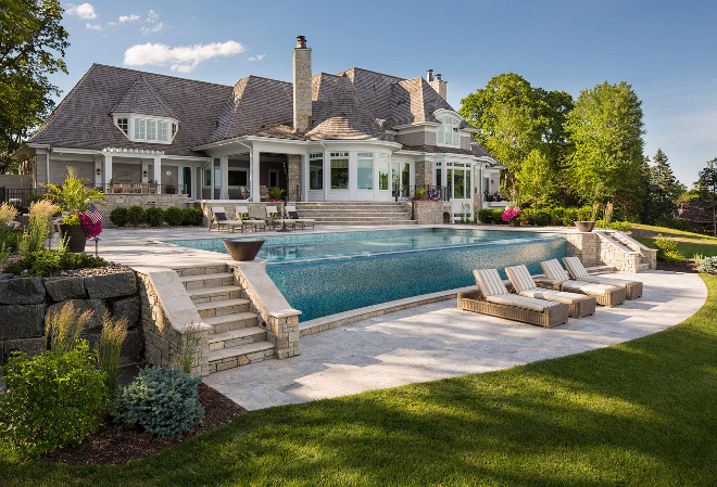 Shingle home backyard. The infinity swimming pool is the centerpiece of this dramatic yard. The upper and lower natural stone patios give depth to the entertaining space. The retaining walls double as a garden bed to bring color into the yard. The fire bowls on either side of the pool light add to the ambiance for evening entertaining. The paving stone is known as Percheron Travertine. The coped railings and treads are Bisque Travertine. Shingle home backyard ideas. #shinglehomes #shinglehome #backyard Southview Design