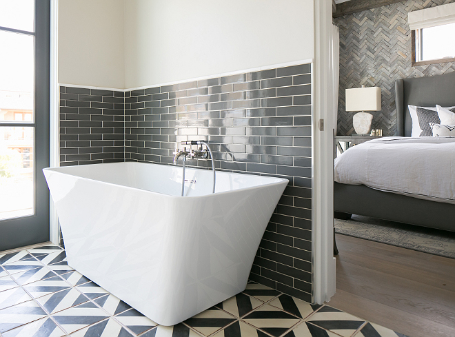 Bathroom. White and gray master bathroom features upper wall painted in a pale grey and lower wall clad in charcoal gray brick tiles lined with a modern freestanding tub and a wall-mount tub filler atop a white and gray geometric tiled floor. The tub is a Tiffany 59" Small Soaking tub from Wyndham. #bathroom Patterson Custom Homes
