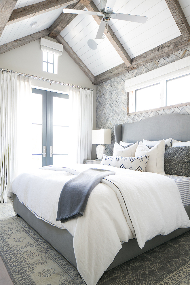 Master Bedroom. This master bedroom feels very original. Gray bedroom boasts a vaulted paneled ceiling adorned with rustic wood trim over a gray herringbone tiled accent wall lined with a gray wingback headboard on bed dressed in white and grey bedding atop a gray rug placed in front of black framed French doors dressed in white drapes. Plank Ceiling Paint Color: Dunn Edwards 2005 Evershield Eggshell. The master bedroom also has vaulted ceilings and French doors to a balcony. Master bedroom wall features white washed reclaimed wood in a herringbone pattern. #masterbedroom #bedroom Patterson Custom Homes 