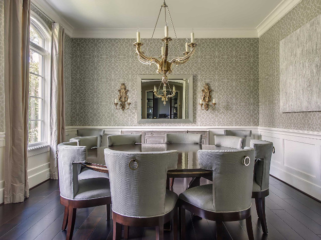 Grey dining room with wainscoting walls. Grey dining room features upper walls clad in gray trellis wallpaper and lower walls clad in wainscoting alongside a chandelier illuminating an expansive round dining table that seats 8 lined with gray velvet dining chairs. #greydiningroom #diningroom #wainscoting #diningroomwainscotingwalls #diningroomwalls Elizabeth Garrett Interiors.