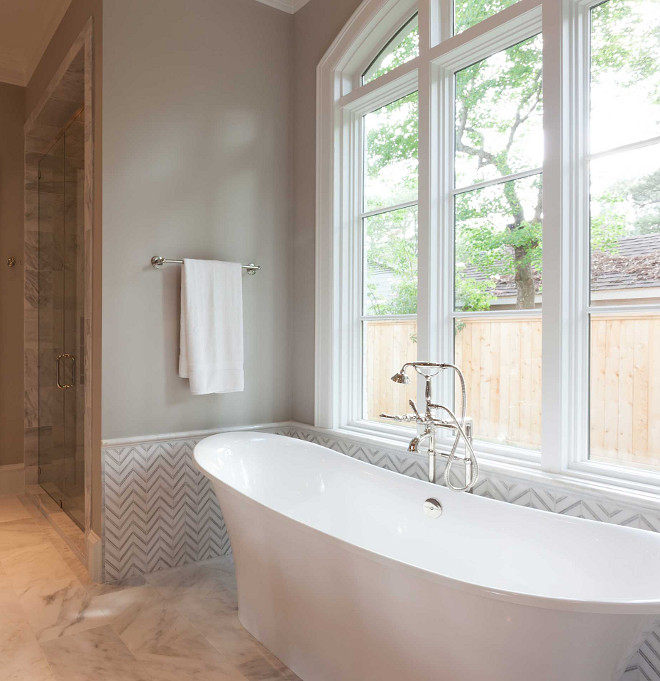 Bathroom features a bathtub nook with upper walls painted gray and lower walls clad in white and gray marble chevron tiles filled with a freestanding tub and a vintage style hand-held tub filler placed under windows. Elizabeth Garrett Interiors