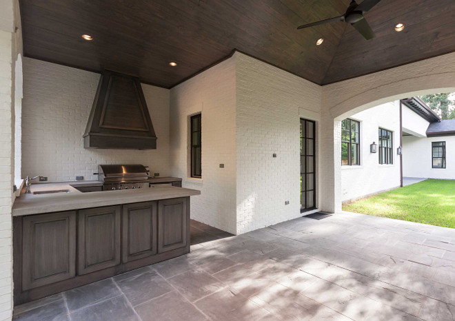 Outdoor Kitchen in Covered Patio. Covered patio boasts a white brick painted wall lined with a walnut stained French vent hood placed over an integrated bbq grill. Outdoor kitchen boasts walnut stained outdoor cabinets paired with concrete countertops fitted with a sink and vintage style faucet.. #outdoorkicthen #outdookitchencabinet Elizabeth Garrett Interiors