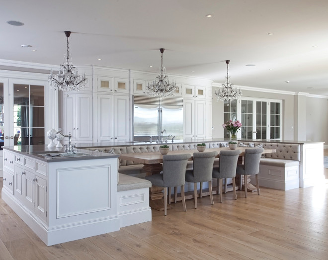 Kitchen island with banquette. We really don't see a kitchen like this every day. This gorgeous kitchen features a very large center island with tons of storage space and an U-shaped dining banquette. The back wall of the kitchen boasts a row of small glass-front cabinets stacked atop pantry cabinets with bi-fold doors flanking stainless steel refrigerators. Also notice the Restoration Hardware Salvaged Wood Trestle Dining Table lined with gray roll back dining chairs. #kitchen #island #banquette Hayburn & Co. 