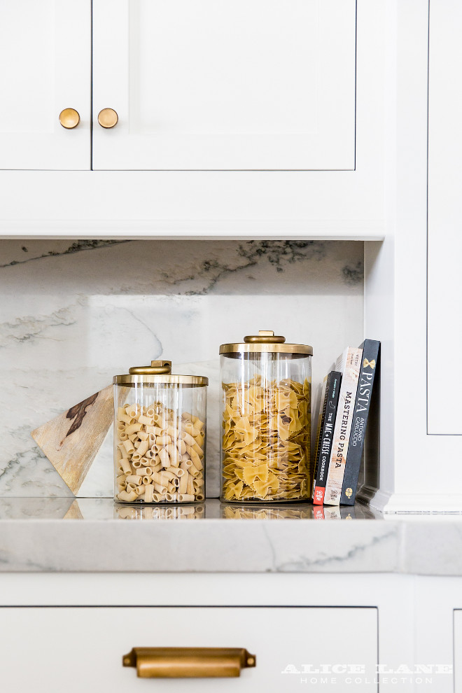 Kitchen Brass Hardware. White Shaker Kitchen Cabinets with Brushed Brass Knobs and Vintage Pulls. kitchen features white shaker cabinets adorned with brushed brass knobs and vintage brass pulls paired with gray and white marble countertops and backsplash lined with glass canisters with gold lids. Alice Lane Home.