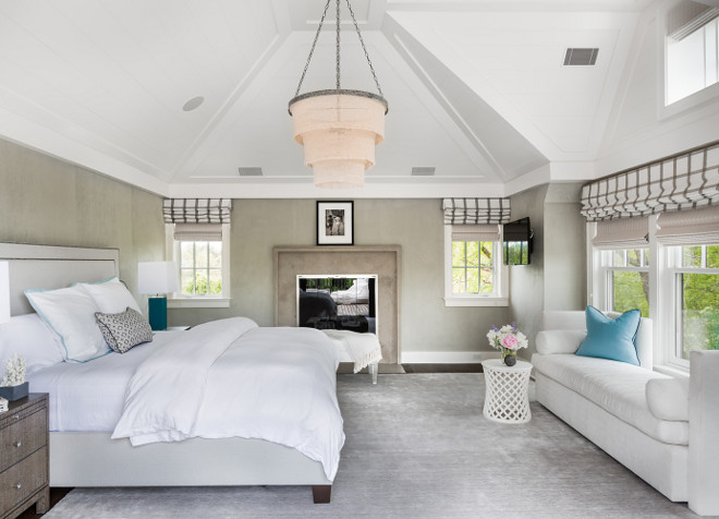 Neutral bedroom with vaulted ceiling accented with a 3 tier linen chandelier illuminating a light gray nailhead bed flanked by wood 2 drawer nightstands and turquoise blue lamps facing a white chaise lounge lined with turquoise blue velvet pillows. Chic bedroom boasts a textured gray wall accented with taupe stone fireplace alongside a white bench flanked by windows dressed in gray striped roman shades. Alisberg Parker Architects.