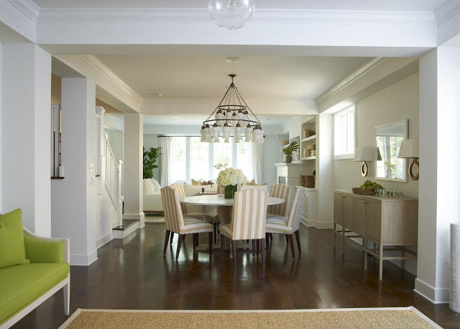 Sophisticated open dining room with white Sara Chandelier over round marble top dining table surrounded by white and tan striped camelback dining chairs. Weathered oak buffet cabinet and white mirror flanked by brass sconces.