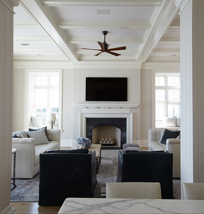 Living room coffered ceiling. The living room features an ivory coffered ceiling, with coffers painted in white accented with a modern wooden ceiling fan. A pair of blue accent chairs facing a shiplap fireplace wall fitted with a flat panel tv over a white fireplace mantel finished with a black stone surround flanked by window seat alcoves.#livingroom #cofferedceiling #tongueandgroove #paneledwalls.