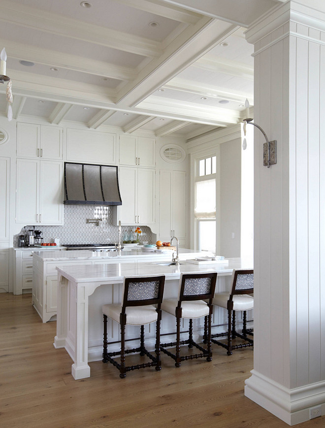White kitchen with coffered ceiling. White kitchen. The kitchen also features double islands and a French hood that stands over a swing arm pot filler and a stainless steel range. One island is designated as a prep station fitted with a sink and gooseneck faucet, while the second kitchen island is designated as the dining table fitted with a small prep sink and white and gray square back counter stools.#whitekitchen #kitchen #cofferedceiling #kitchencofferedceiling TS Adams Studio Architects. Laura Allyson Interiors.