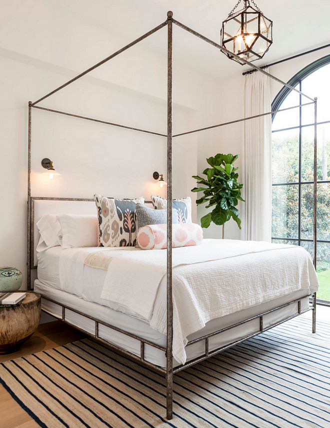 Bedroom with metal canopy bed, Oly Studio Marco Bed, dressed in soft white bedding and pink and gray ikat pillows as well as a pink ikat bolster pillow placed atop a beige and navy striped rug illuminated by glass vintage barn wall sconces and a Suzanne Kasler Morris Lantern. Coats Homes