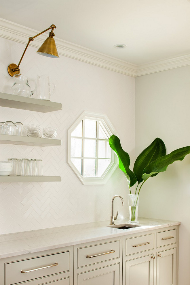 Floating open shelves. Pale Gray Floating Open Shelves. Floating pale gray shelves are lit by Boston Functional LIbrary Wall Lights mounted on white chevron backsplash tiles framing an octagon window located over a small prep sink fitted with a polished nickel gooseneck faucet fixed in Bianco Macabus Quartzite countertops positioned atop gray shaker cabinets finished with polished nickel pulls. #palegray #openshelves #floatingshelves Revival Construction