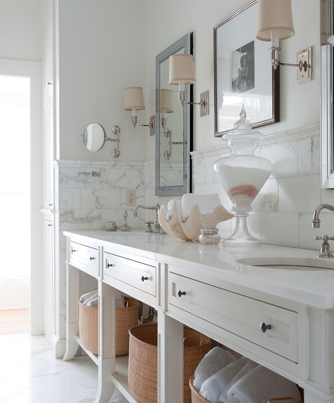 White bathroom features a white dual washstand fitted with three drawers and a shelf lined with pale pink wicker baskets. A white dual bath vanity is fitted with oval sinks and vintage cross handle faucets accented with a pale pink clam shell lining a half tiled marble wall finished with marble chair rails. A white master bath vanity stands under beveled mirrors illuminated by polished nickel sconces dressed in pale pink sconces. #bathroom #whitebathroom #bathrooms #bathroomideas #bathroomreno #bathroomwashstand #washstand Smith Hanes 