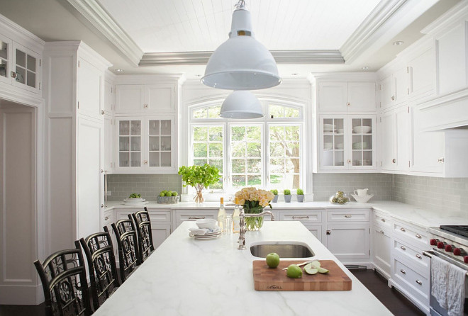 A kitchen doorway is fitted with glass display cabinets. A kitchen doorway is fitted with glass display cabinets leading to a beautiful kitchen filled with white shaker cabinets paired with white marble countertops and a gray subway tile backsplash. A pair of white industrial pendants hang over a white center island fitted with a sink lined with brown bamboo counter stools by Redegg Indochine Bar Stools. A stainless steel sink stands under arched windows flanked by glass door cabinets. #kitchen #doorway Dibico Construction