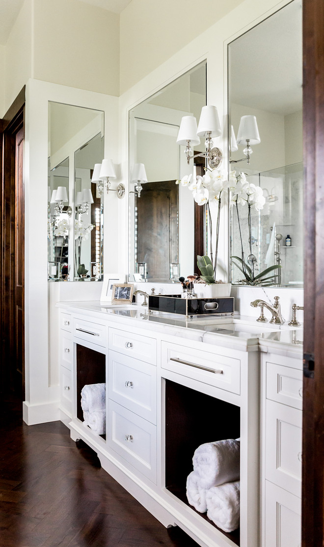  White Dual Bath Vanity with Dark Wood Herringbone Floor: Exquisite master bathroom features a white double washstand fitted with open bins lined with rolled up towels topped with white beveled marble surrounded by four framed mirrors illuminated by Robert Abbey One Light Crystal Wall Sconces alongside a dark stained wood herringbone floor. bathroom-cabinet-white-dual-bath-vanity-with-dark-wood-herringbone-floor-gallery