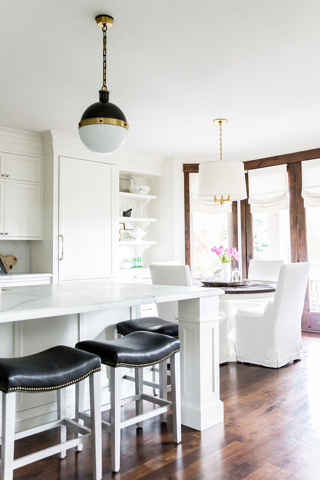 Kitchen lighting. Stunning kitchen features a pair of Hicks Pendants and a drum chandelier with brass chain, Dalston Hanging Shade, hangs over a white dining table accented with a stained wood top surrounded by white slipper dining chairs. Stunning kitchen features a pair of Hicks Pendants illuminate a long white center island fitted with a farmhouse sink and deck mount vintage faucet lined with black leather saddle seat counter stools. A drum chandelier with brass chain, Dalston Hanging Shade, hangs over a white dining table accented with a stained wood top surrounded by white slipper dining chairs. #kitchenlighting #HicksPendants #DalstonHangingShade LIV Design Collective.